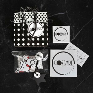 May include: A black and white polka dot gift bag with a black handle, a small white box, stickers, and tags. The stickers and tags feature the text "GOZEMRE atelier" in a circular design. A clear bag contains a stick figure design.