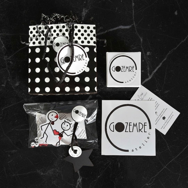 May include: A black and white polka dot gift bag with a black handle, a small white box, stickers, and tags. The stickers and tags feature the text "GOZEMRE atelier" in a circular design. A clear bag contains a stick figure design.