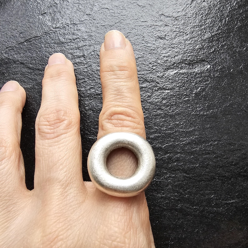 May include: A silver ring with a thick, rounded band is worn on a finger. The ring has a smooth, matte finish and a circular design. The ring is a statement piece of jewellery.