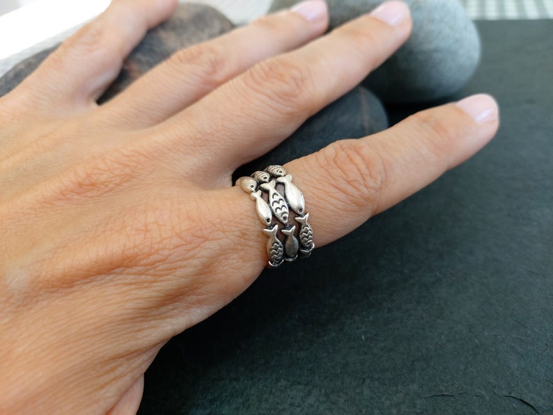 May include: A silver ring with three bands of fish designs. The ring is on a person's finger.