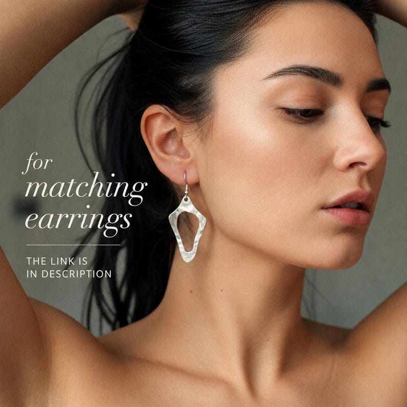 May include: A silver-toned drop earring with a textured, hammered finish and an irregular, elongated diamond shape featuring a central cutout. The image includes the text: for matching earrings THE LINK IS IN DESCRIPTION.