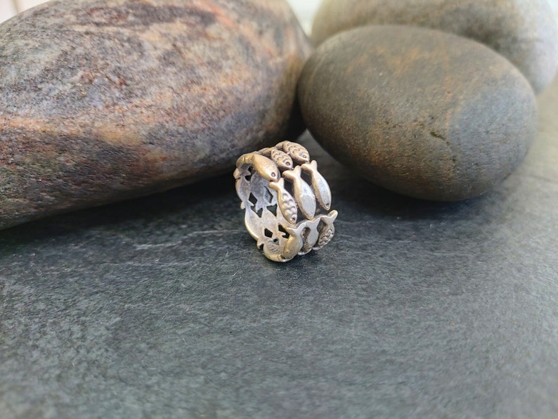 May include: A silver ring with a design of multiple fish, each facing the same direction, encircling the band. The ring is on a dark gray surface.