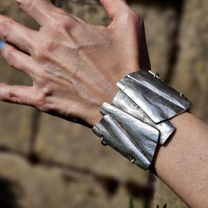 May include: A silver-toned cuff bracelet with a layered design. The bracelet features rectangular plates with a textured, wood-grain appearance. The metal has a brushed finish, and the bracelet is secured with small hinges.