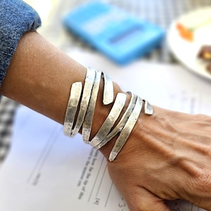 May include: A silver-toned cuff bracelet with a textured, hammered finish. The bracelet wraps around the wrist with multiple overlapping bands. The jewelry is worn on a person's arm.
