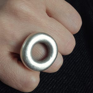 May include: A silver ring with a circular design. The ring has a smooth, matte finish and a thick band. The ring is worn on a finger, and the design is a solid, rounded shape with a hole in the centre.