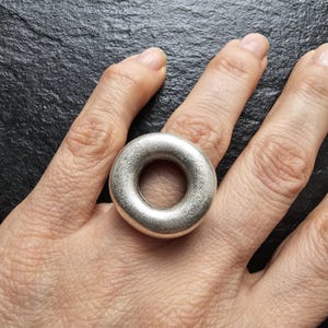 May include: A silver ring with a circular design. The ring is thick and has a smooth, matte finish. The ring is worn on a finger, showcasing its simple, yet bold design. The ring's shape is a perfect circle with a hole in the centre.