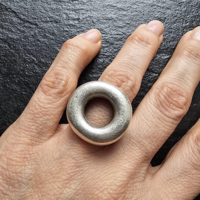 May include: A silver ring with a circular design. The ring is thick and has a smooth, matte finish. The ring is worn on a finger, showcasing its simple, yet bold design. The ring's shape is a perfect circle with a hole in the centre.