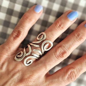 May include: A silver ring with a swirling, abstract design. The ring is worn on the index finger of a hand with light blue nail polish.
