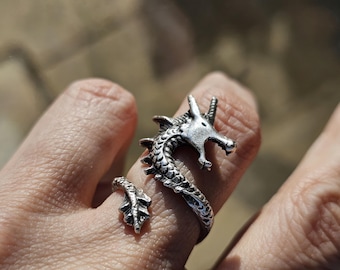 Op de afbeelding: Een zilveren drakenring met een opgerolde staart en een open mond. De ring wordt aan een vinger gedragen.
