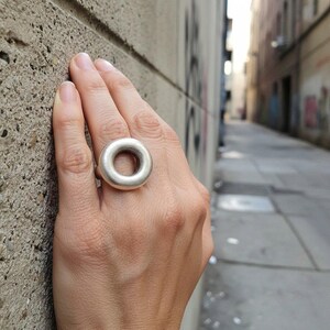 May include: A silver ring with a circular design. The ring is worn on a hand, with a textured concrete wall in the background. The ring's shape is a solid circle with a hole in the centre, creating a modern, minimalist aesthetic.