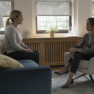 May include: A blue sofa with gold pillows, a light gray armchair, a white table lamp, and a wooden radiator cover. Two individuals are seated in the room, one on the sofa wearing a light gray long-sleeve shirt and one in the armchair wearing a gray long-sleeve shirt and dark trousers.