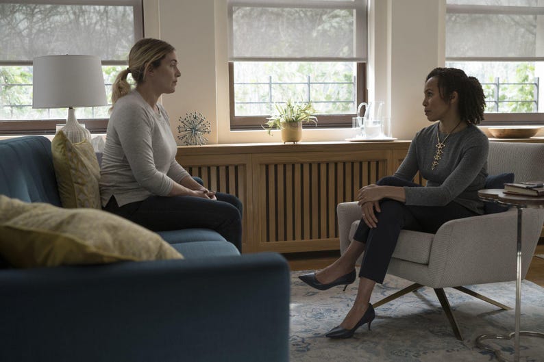 May include: A blue sofa with gold pillows, a light gray armchair, a white table lamp, and a wooden radiator cover. Two individuals are seated in the room, one on the sofa wearing a light gray long-sleeve shirt and one in the armchair wearing a gray long-sleeve shirt and dark trousers.
