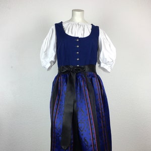 Könnte beinhalten: Ein blaues Dirndl mit schwarzem Band und einer weißen Bluse. Der Rock hat ein schwarz-blaues Muster mit roten Akzenten.