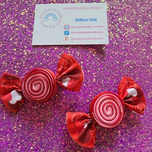 May include: Two red and white candy-shaped hair bows with a spiral design. The bows have a red and white heart pattern on the ends. A business card with social media handles is in the background.
