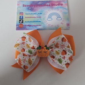 May include: A hair bow featuring solid orange ribbon layered with white ribbon printed with colourful autumn leaves and acorns. A small orange pumpkin charm with a unicorn horn and closed eyes is centred on the bow. A business card for ScoobyDoodles Studio is visible above the bow.