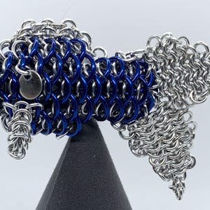 May include: A blue and silver chainmail fish sculpture. The fish is made of small metal rings that are linked together to create a scale-like effect. The fish is displayed on a black stand.