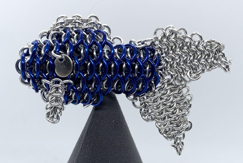 May include: A blue and silver chainmail fish sculpture. The fish is made of small metal rings that are linked together to create a scale-like effect. The fish is displayed on a black stand.