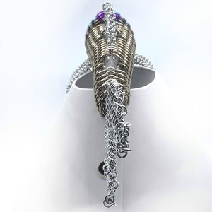 May include: A silver chainmaille necklace with a gold chainmaille pendant. The pendant is decorated with purple and blue beads.