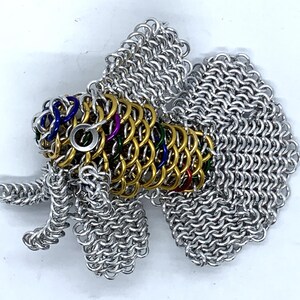 May include: A silver and gold chainmail fish sculpture with colorful accents. The fish has a large tail fin and a smaller dorsal fin. The sculpture is made of small metal rings that are linked together.