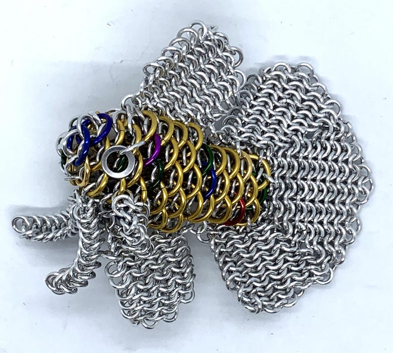 May include: A silver and gold chainmail fish sculpture with colorful accents. The fish has a large tail fin and a smaller dorsal fin. The sculpture is made of small metal rings that are linked together.