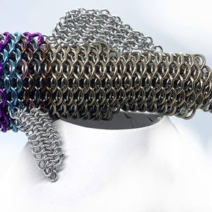 May include: A silver, purple, and blue chainmail fish sculpture. The fish has a long, slender body with a pointed head and a forked tail. The chainmail is made of small, interlocking metal rings.