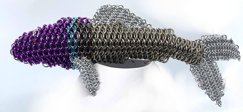 May include: A silver, purple, and blue chainmail fish sculpture. The fish has a long, slender body with a pointed head and a forked tail. The chainmail is made of small, interlocking metal rings.