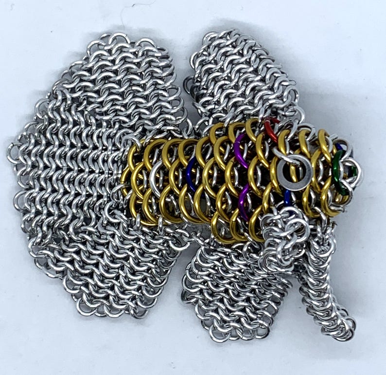 May include: A silver and gold chainmail fish sculpture with colorful accents. The fish has a long, flowing tail and fins.