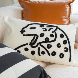 Könnte beinhalten: Ein weißes Sofa mit drei Kissen. Die Kissen sind ein schwarz-weiß gestreiftes Kissen, ein weißes Kissen mit einer schwarzen Umrandung eines Leoparden und ein braunes Lederkissen.