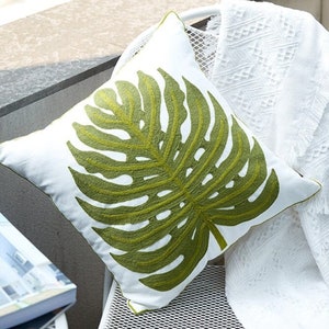 Puede incluir: Cojín cuadrado blanco con un gran diseño de hoja de monstera verde bordada. El cojín tiene un ribete verde y está colocado sobre una silla blanca con una manta blanca. Un artículo decorativo.