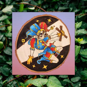 May include: A circular wood slice featuring a pyrography and painted illustration of a jester in a blue and red costume. The jester holds a wooden marionette cross control. The background is divided diagonally into black and light wood sections with stars.