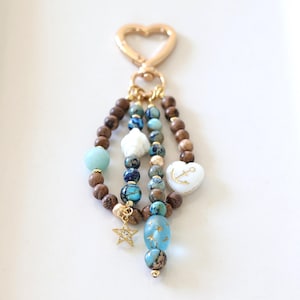 May include: A keychain featuring a gold-toned heart-shaped clasp attached to three strands of beads. The strands include brown wooden beads, blue and turquoise marbled stones, a white heart charm with a gold anchor, and a gold star charm.
