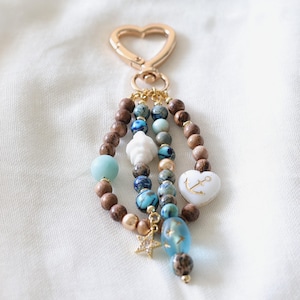 May include: A beaded keychain with a gold heart-shaped clasp. The keychain features various beads in shades of brown, blue, and white, including a seashell, a heart with an anchor, and a star charm. The beads are strung together to create a decorative accessory.