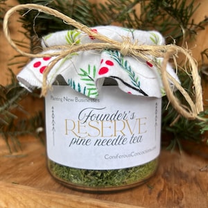 Peut inclure: Un pot en verre de thé aux aiguilles de pin Founder's Reserve. Le pot est scellé avec un couvercle en tissu blanc orné de baies rouges et de feuilles vertes, noué avec de la ficelle. L'étiquette indique "Founder's Reserve pine needle tea".