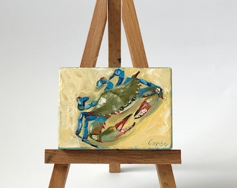 May include: A small, square oil painting of a blue crab with green and red accents, displayed on a miniature wooden easel. The crab is painted on a textured, light yellow background. The artist's signature is visible in the lower right corner.