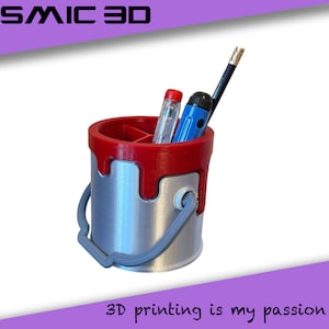 May include: A 3D-printed pen holder in the shape of a paint bucket. The top rim and paint drips are red, the bucket is silver, and the handle is grey. It holds a pencil, a blue pen, and a red pen. The text "COSMIC 3D" is at the top.