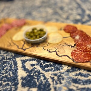 May include: A wooden serving tray with a blue resin design. The tray is filled with sliced salami, potato chips, and a small bowl of green olives.