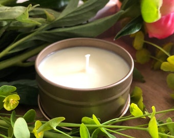Puede incluir: Una vela pequeña de lata marrón con una parte superior de cera blanca y una sola mecha. La vela está rodeada de follaje verde y flores rosas.