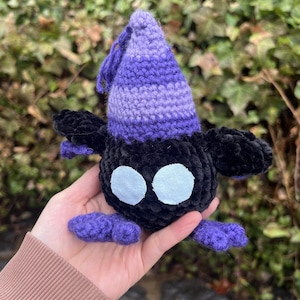 Puede incluir: Una criatura de ganchillo hecha a mano en negro y morado. El peluche tiene un gorro puntiagudo con rayas moradas y lavanda, ojos blancos grandes y pequeños pies y alas moradas. El juguete se sostiene en una mano.