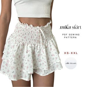 May include: A white floral print skirt with a ruffled hem and smocked waistband. It has a drawstring tie. The text "mika skirt" is displayed, along with "PDF Sewing Pattern" and size range "XS-XXL".