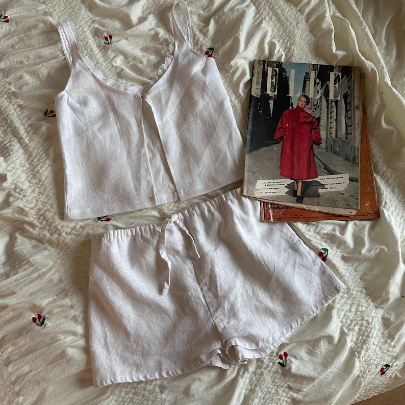 May include: A white linen pajama set with a lace trim on the top and a drawstring on the shorts. The set is laid out on a white bedspread with a magazine titled "ELLE" on top.