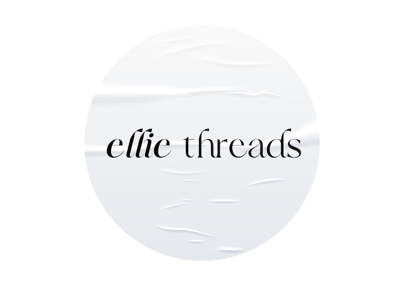 May include: A white, circular sticker with the words "ellie threads" in black script. The sticker has a textured, wrinkled appearance, suggesting it has been applied to a surface. The design is simple and elegant.