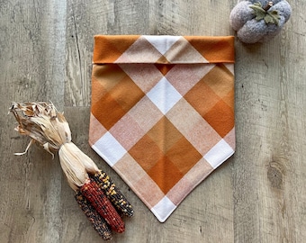 May include: Orange and white plaid bandana with a folded edge. The bandana is on a wood background with a small gray pumpkin and corn husks.