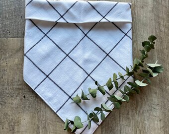 May include: White cotton bandana with a black and white plaid pattern. The bandana has a folded edge and is lying on a wooden surface.