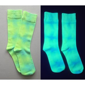 May include: A pair of crew-length socks featuring a tie-dye pattern in shades of neon yellow and light blue. The socks are shown in two states: one pair in normal lighting and another pair glowing in the dark with a bright, luminous green appearance.