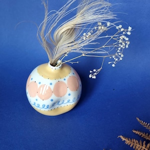 Könnte beinhalten: Eine kleine, runde Keramikvase mit gelbem Boden und weißem Körper, verziert mit rosa Kreisen und blauen Punkten. Die Vase enthält eine Feder und zarte weiße Blumen vor blauem Hintergrund. Die Vase ist ein dekorativer Gegenstand.