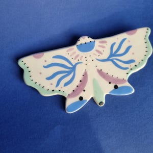 Puede incluir: Una mariposa de cerámica con base blanca, adornada con detalles pintados en azul, morado y verde. Las alas presentan patrones azules estilizados y pequeños puntos negros. La mariposa está sobre un fondo azul liso.
