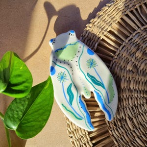 Könnte beinhalten: Eine weiße, froschförmige Keramikschale mit blauen, grünen und türkisfarbenen floralen Designs. Die Schale hat eine gebogene Form mit einem Ausschnitt am Boden. Die Augen des Frosches sind blau.