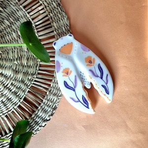 Può includere: Un piatto da portata in ceramica bianca a forma di chela di aragosta, decorato con motivi floreali arancioni e viola. Il piatto è appoggiato su un sottobicchiere intrecciato e su uno sfondo color pesca. Il piatto misura circa 20 cm di lunghezza.