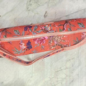 May include: A cylindrical yoga mat bag in a vibrant orange hue, adorned with a floral pattern in shades of pink, purple, and green. The bag features a white zipper, a matching strap, and gold-toned hardware. The bag is designed to carry a yoga mat.