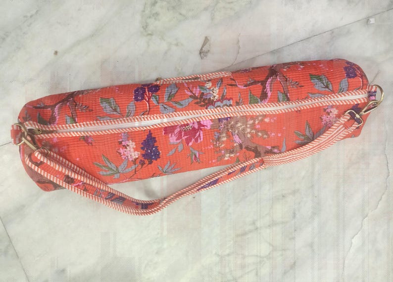May include: A cylindrical yoga mat bag in a vibrant orange hue, adorned with a floral pattern in shades of pink, purple, and green. The bag features a white zipper, a matching strap, and gold-toned hardware. The bag is designed to carry a yoga mat.